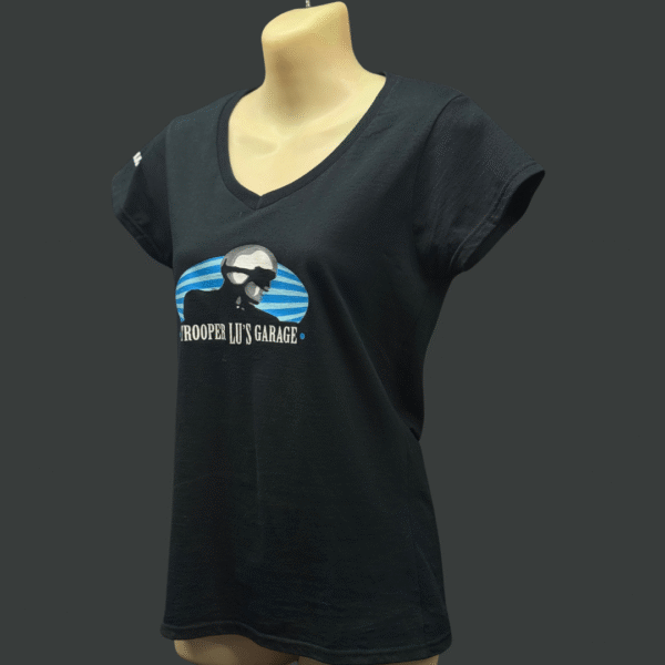 The TROOPER LU T-SHIRT - WOMEN'S is a black V-neck displayed on a mannequin, featuring a graphic of a bearded man with sunglasses and cap, plus "TROOPER LU'S GARAGE" text on the front.