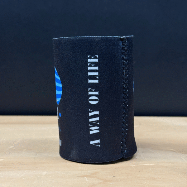 A TROOPER LU'S GARAGE STUBBY COOLER, black with vertical white "A WAY OF LIFE" text, stands on a wooden surface against a dark background.
