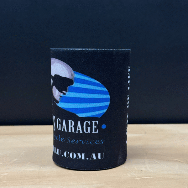 The TROOPER LU'S GARAGE STUBBY COOLER is a black cylindrical beverage holder with a blue and white stylized skull logo, "GARAGE Motorcycle Services" text, and a partial website address.