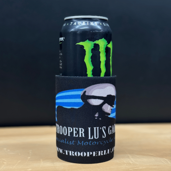A Monster Energy drink can in a TROOPER LU'S GARAGE STUBBY COOLER on a wooden surface with a black background.
