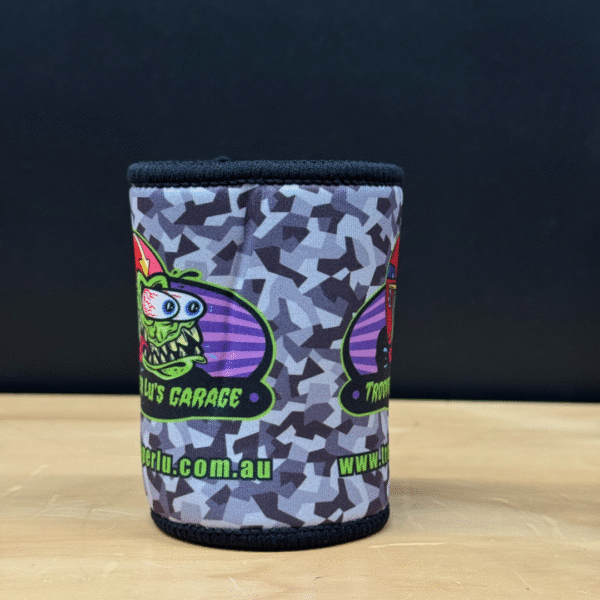 A TROOPER LU'S GARAGE STUBBY COOLER with a grey and black pattern, cartoon monster, and "Troy’s Garage" plus "troyberlu.com.au" text on a wooden surface against a black background.