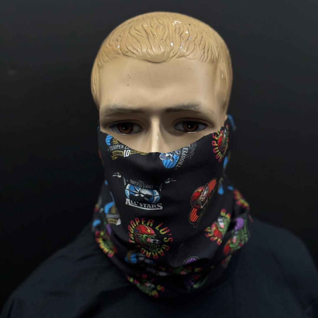 A mannequin with blonde hair models the HEADSKINZ TROOPER LU'S NECKWARMER, a black neck gaiter adorned with colorful logos and text, covering the lower half of its face against a plain dark background.