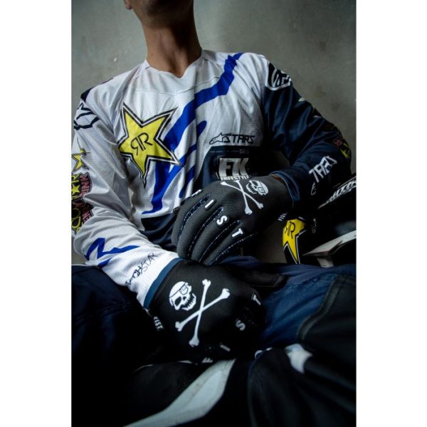 A person sits with a helmet in their lap wearing Fist Rodger Gloves, black with skull and crossbones, along with a white and blue motocross jersey adorned with star logos.